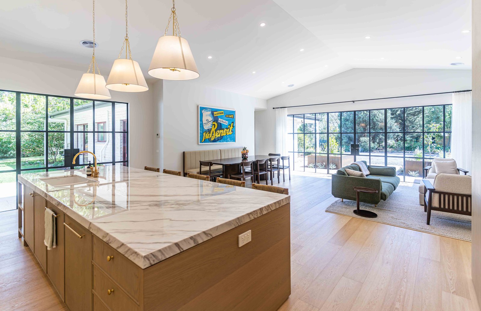 Spacious modern kitchen and living area with a large marble island, dining table, and floor-to-ceiling glass doors opening to a garden.