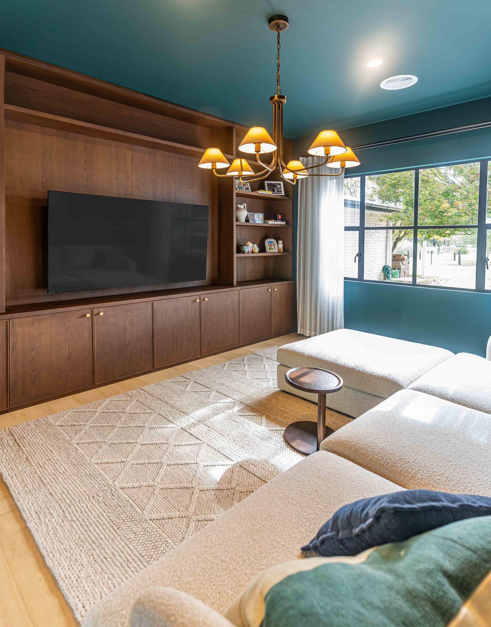 Cozy living room with beige sectional sofa, wooden side table, large TV on wood-paneled wall, and chandelier above.