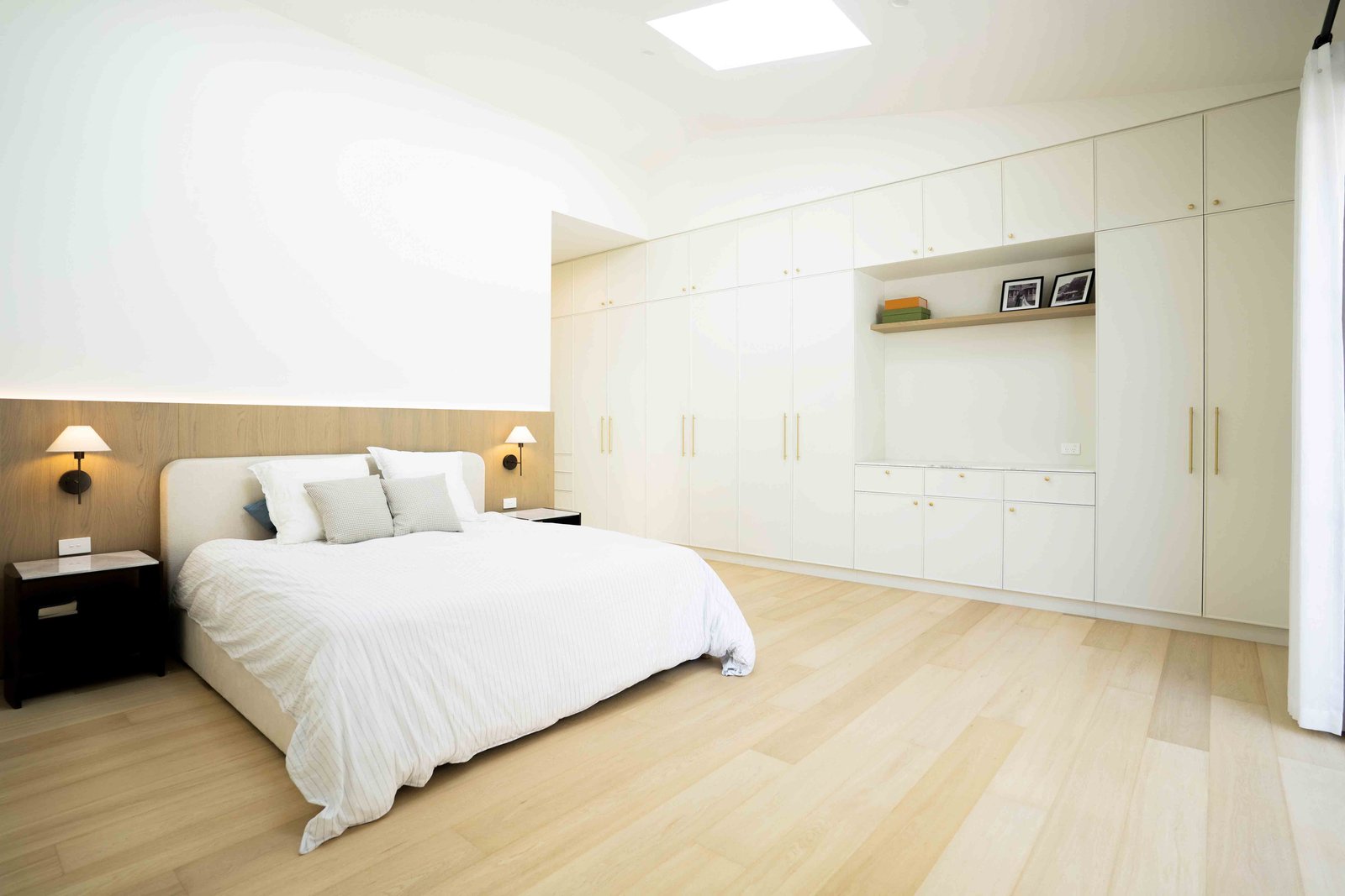 Bright modern bedroom with a large bed, white bedding, wooden floor, built-in white wardrobes, and two wall lamps.