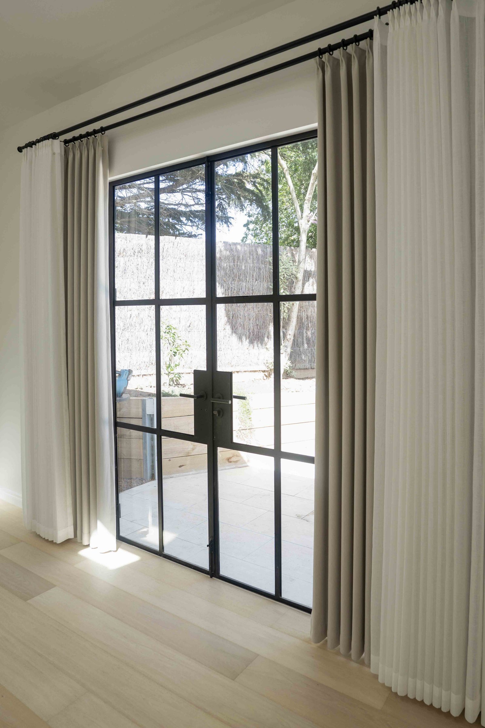 Modern black-framed glass doors with white and beige pleated curtains on a light wooden floor.