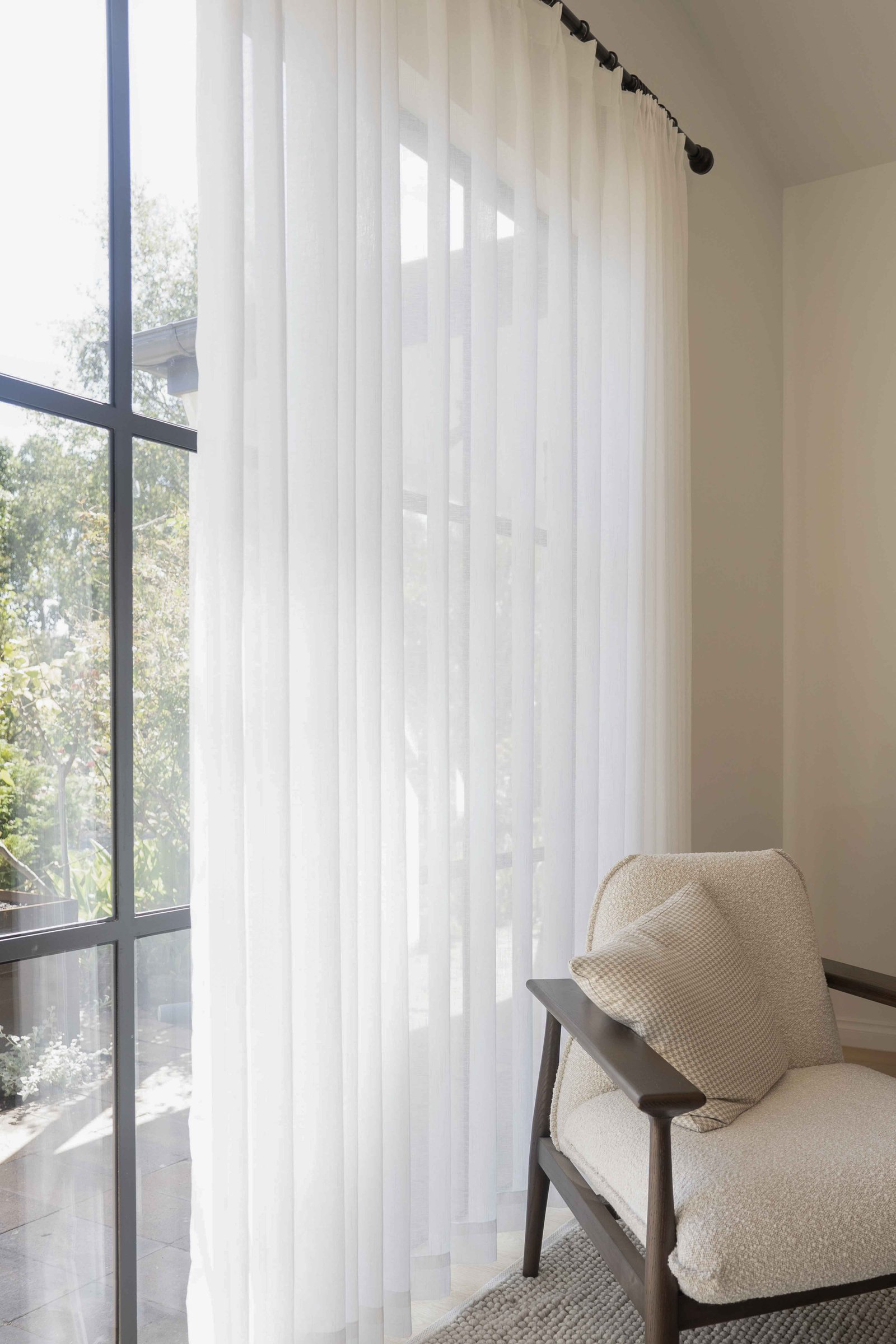 Cozy cushioned armchair with a patterned pillow beside large window with GETC sheer white curtains.