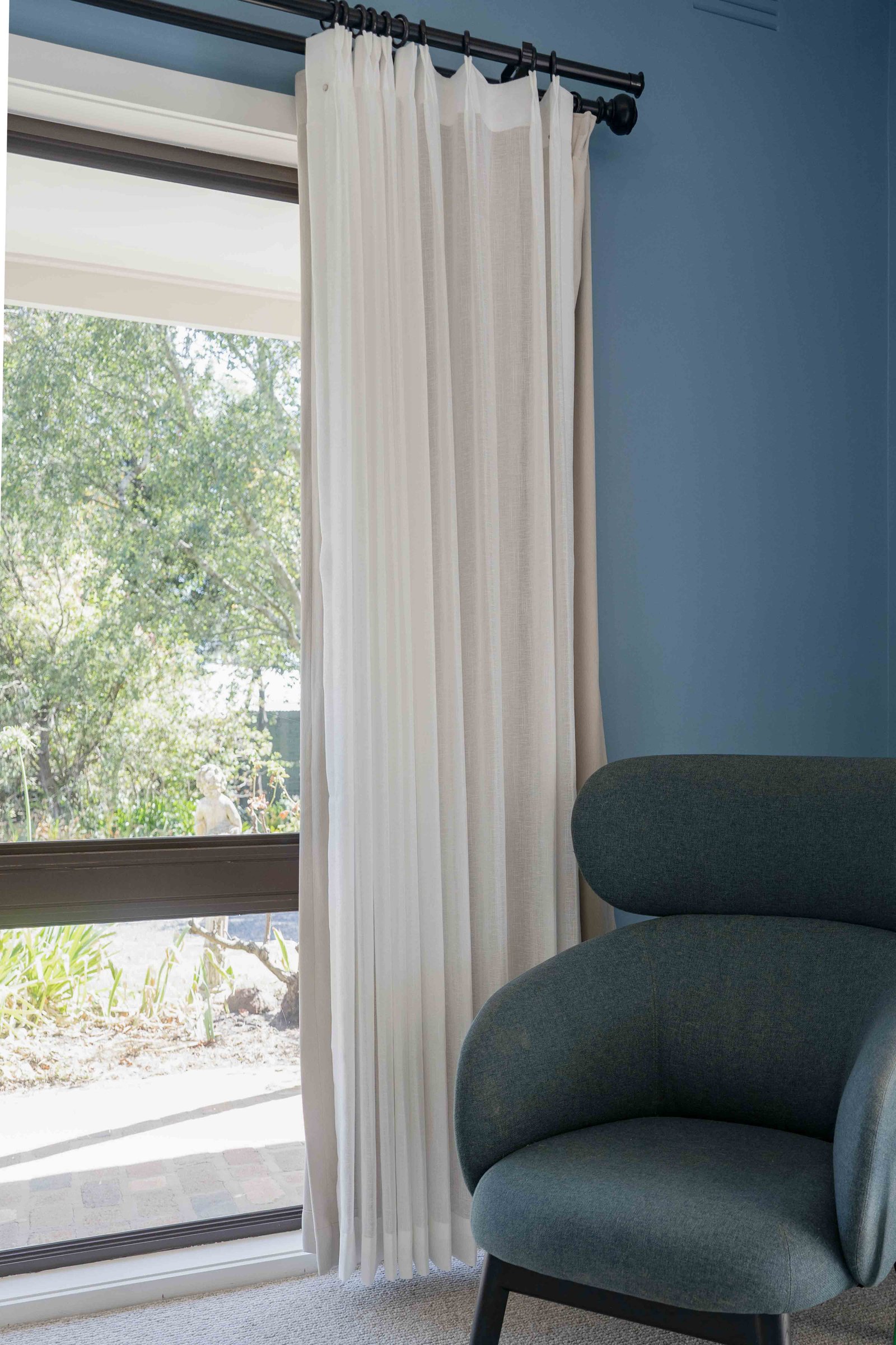 Cream-colored and sheer white curtains hanging on a black rod next to a large window with a view of green trees, and a dark blue upholstered armchair nearby.