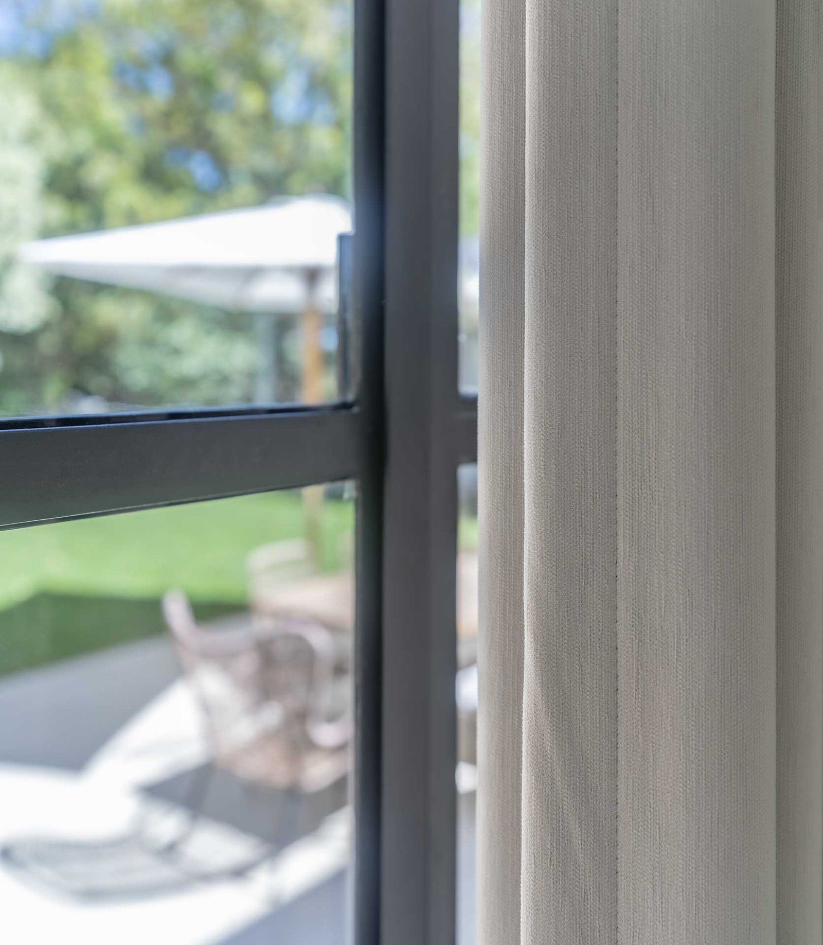 Close-up of a beige curtain beside a black metal-framed window with a blurred outdoor patio and green garden in the background.