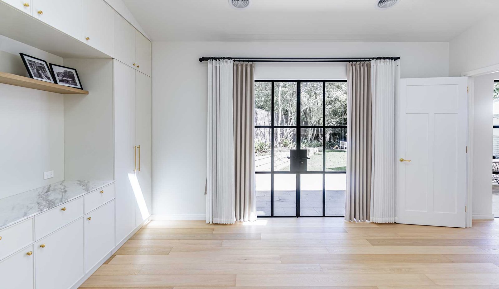 Bright room with light wood flooring, white built-in cabinets with marble countertop, and black-framed glass French doors opening to a garden.